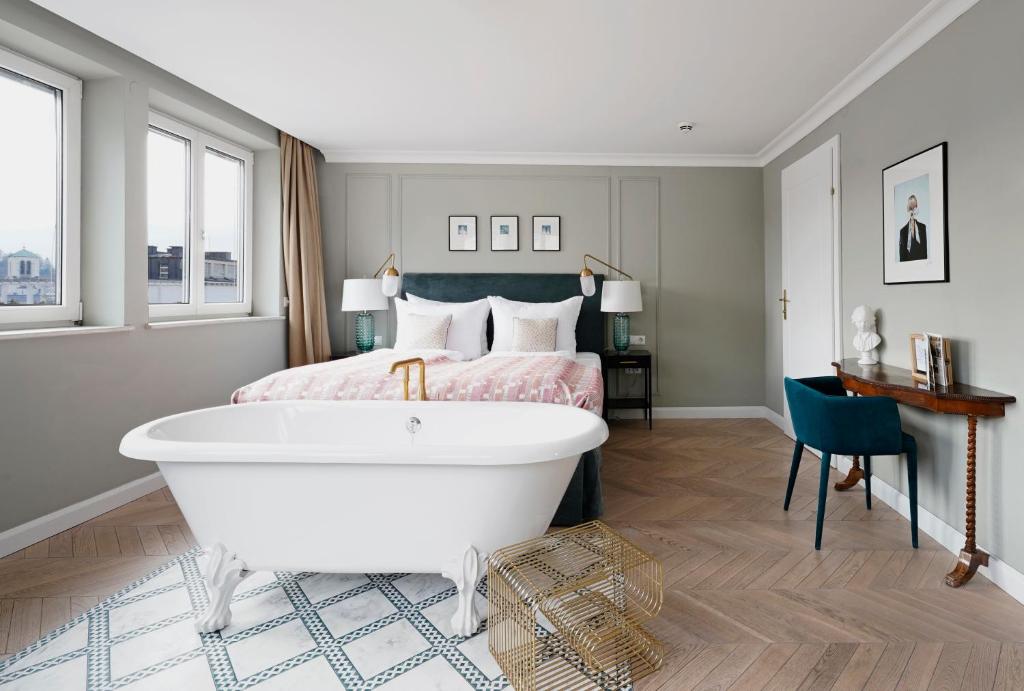Unique Boutique Hotels in Salzburg 15 A modern bedroom reminiscent of boutique hotels in Salzburg, featuring a freestanding white bathtub on patterned tile, a double bed with pink bedding, two lamps, large windows, a chair, a small desk, and minimalist decor.