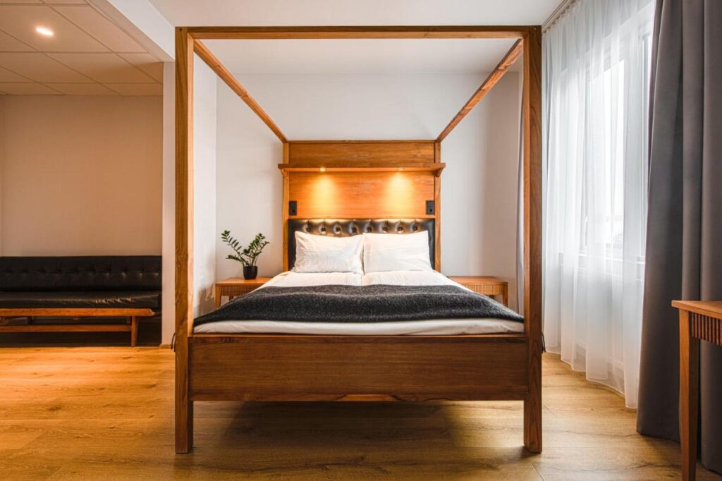 Where to Stay in Reykjavík: Top Guide 22 A modern bedroom, perfect for those wondering where to stay in Reykjavik, features a wooden four-poster bed, white bedding, a dark blanket, wood floors, black sofa, side table with a plant, and large windows with sheer curtains filtering natural light.