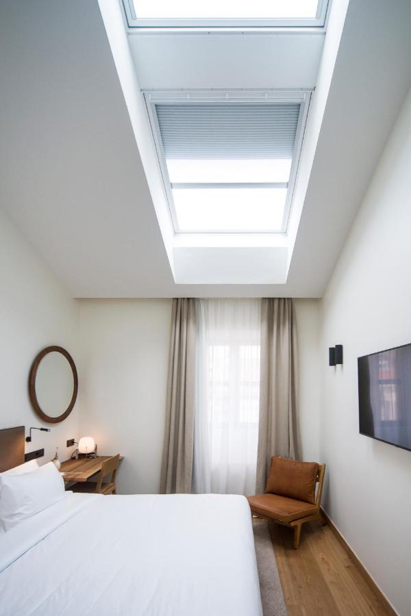 Best Boutique Hotels in San Sebastian 8 Minimalist bedroom inspired by boutique hotels in San Sebastian, featuring a large skylight, white walls, a bed with white linens, a wooden desk with lamp, round mirror, brown chair, sheer curtains, and a wall-mounted TV.