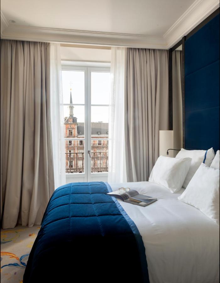 Where to Stay in Madrid: 14 Best Hotels 8 A neatly made bed with white sheets and a blue comforter sits beside a window overlooking historic buildings and a church spire—an inviting scene for those searching where to stay in Madrid first time. An open book rests on the bed.