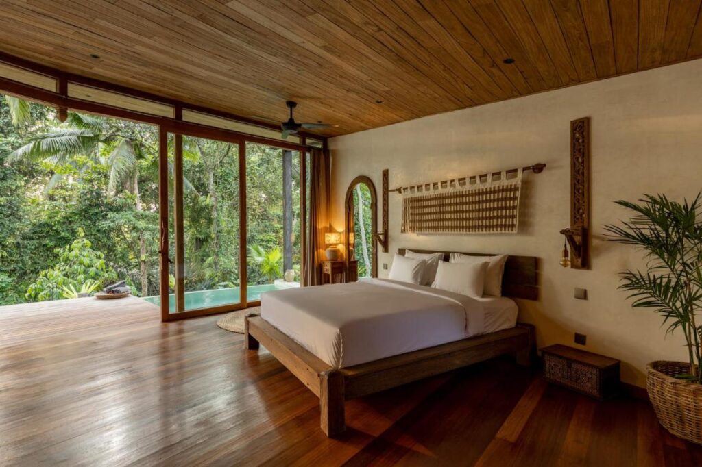 A spacious bedroom with a wooden floor and ceiling, a large bed with white linens, tropical decor, and floor-to-ceiling windows opening to a deck overlooking lush greenery and a small pool—perfect for those seeking the best family villas in Bali.