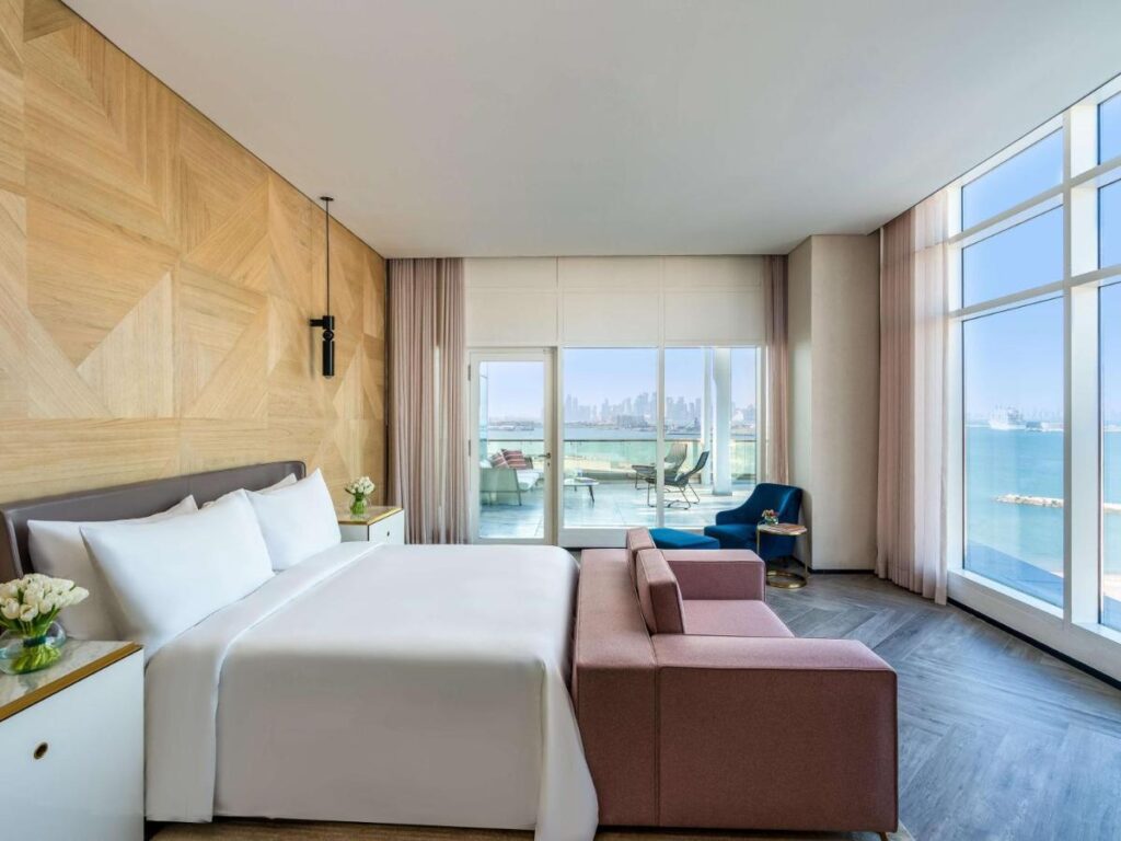 Where to Stay in Doha, Qatar: Transit, City & Beach Hotels 25 Modern hotel room with a large bed, pink sofa, and blue chair by floor-to-ceiling windows offers sea and city views—ideal for those searching where to stay in Doha. A balcony with outdoor seating is visible through glass doors.