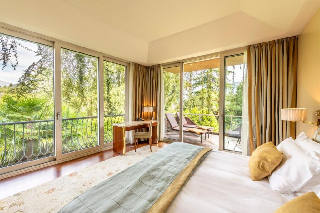 Bright bedroom with large windows and a door leading to a balcony with lounge chairs. Similar to the best hotels in Lugano, this room features white linens, yellow pillows, a desk with chair, and views of green trees outside.