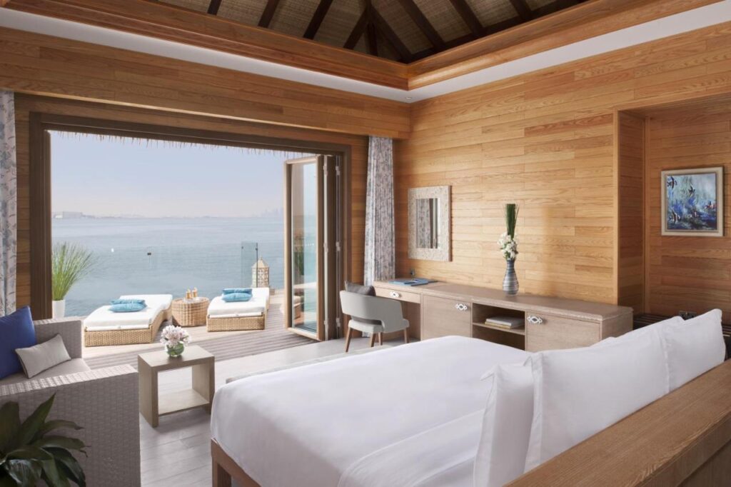 Where to Stay in Doha, Qatar: Transit, City & Beach Hotels 14 A modern, sunlit bedroom with wood-paneled walls, a large bed, desk, and sitting area opens to a patio with lounge chairs overlooking a calm ocean and distant skyline—an ideal choice for where to stay in Doha.