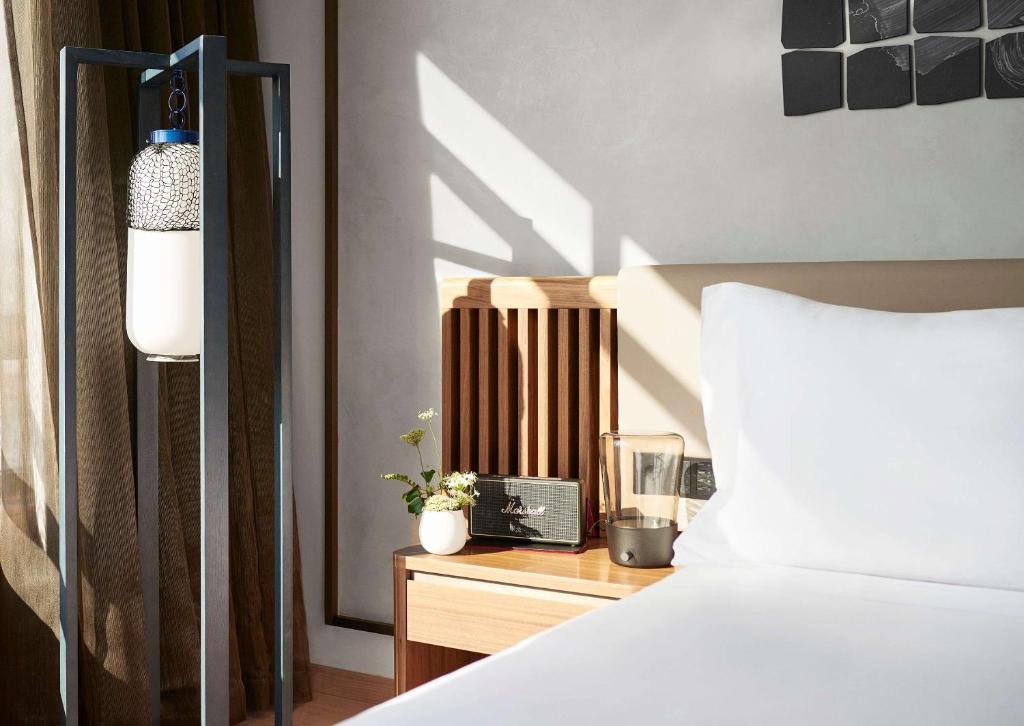 Top Barcelona Hotels with Rooftop Pool 41 A sunlit modern bedroom reminiscent of Barcelona Hotels with Rooftop Pool features a neatly made bed with white linens, a wooden headboard, a nightstand with a glass of water, flowers, and a unique floor lamp beside brown curtains.