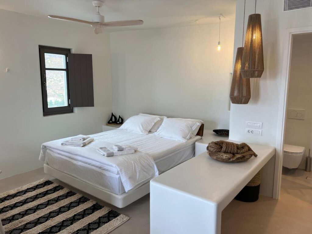 Authentic Hotels in Sikinos, Cyclades 2 A modern, minimalist bedroom in one of the top hotels in Sikinos, featuring a white double bed, neatly folded towels, hanging wicker lamps, a black and white rug, and abundant natural light. A small adjacent bathroom is partially visible.