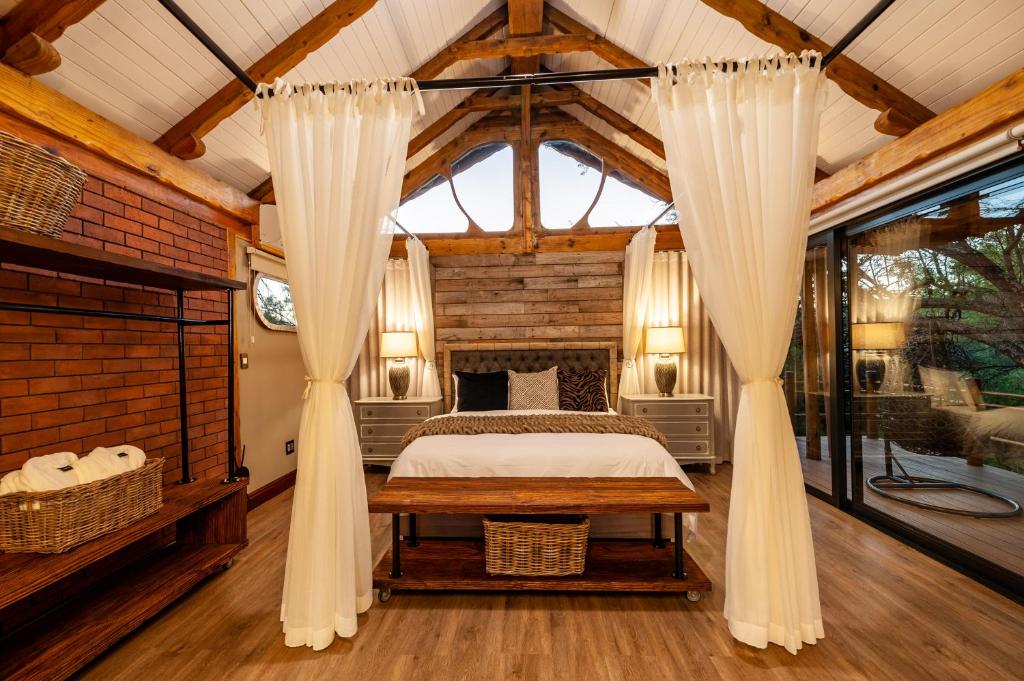 A cozy bedroom with a wooden ceiling and exposed beams—typical of game lodges in Western Cape—features a large bed with white linens, brick walls, sheer curtains, and a sliding glass door leading to a deck with outdoor seating.