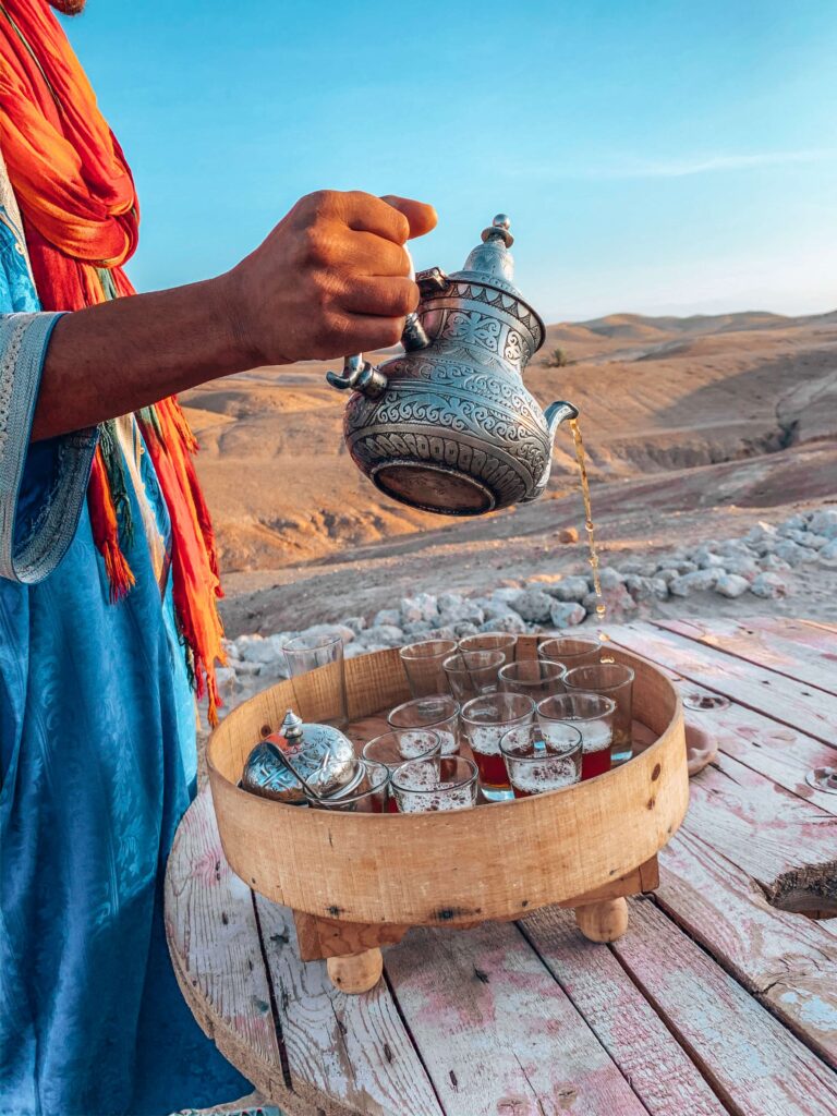 8 SPECTACULAR Agafay Desert Camps 9 A person in a blue robe with an orange scarf pours tea from an ornate metal teapot into small glasses on a wooden tray, set on a rustic table outdoors, capturing the essence of agafay desert camps in the background.