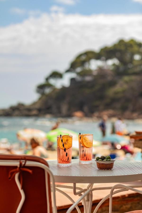 Two pink drinks with lemon slices and a bowl of olives sit on a white table by the seaside—an inviting scene reminiscent of afternoons at boutique hotels in Costa Brava, with vibrant umbrellas and greenery under a partly cloudy sky.