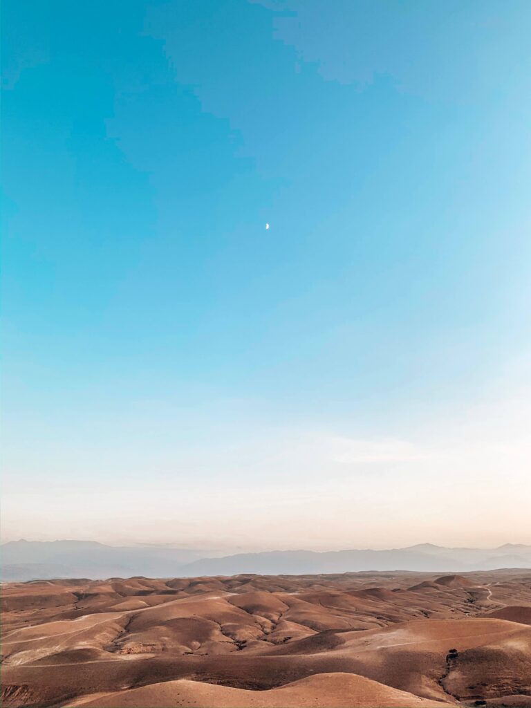 8 SPECTACULAR Agafay Desert Camps 44 Vast desert landscape with rolling sandy hills under a clear blue sky, reminiscent of Agafay Desert Camps. A small white moon is visible high above the horizon, adding to the calm and expansive atmosphere.