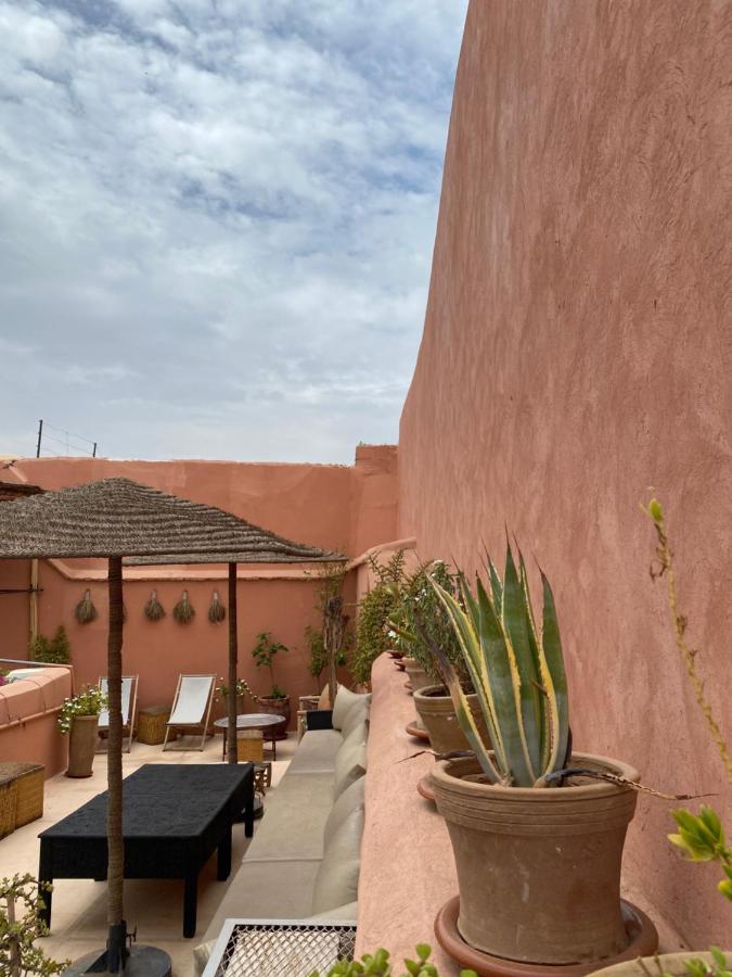 12 Charming Budget Riads in Marrakech 15 A rooftop terrace at one of the budget riads in Marrakech features potted plants, lounge chairs, a black table, and a straw pergola. Pink walls and a cloudy sky complete the relaxed, Mediterranean vibe.