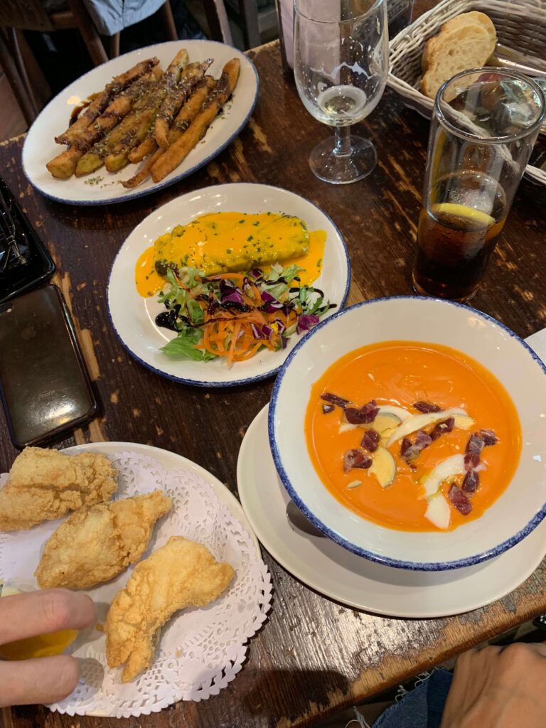 1 day in Seville Spain: My Favorite City 21 A rustic table set with bowls of tomato soup topped with cream and ham, salad, omelette, fried finger foods, bread, and drinks captures a cozy meal—perfect for savoring during 1 day in Seville, Spain at a welcoming restaurant.
