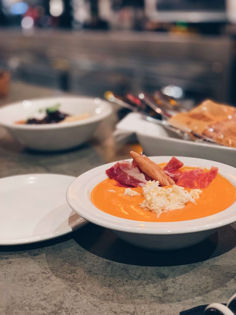 1 day in Seville Spain: My Favorite City 16 A white bowl filled with orange creamy soup, garnished with shredded egg, ham slices, and a breadstick sits on a grey countertop—perfect for savoring during 1 day in Seville, Spain—with another dish and a plate blurred in the background.