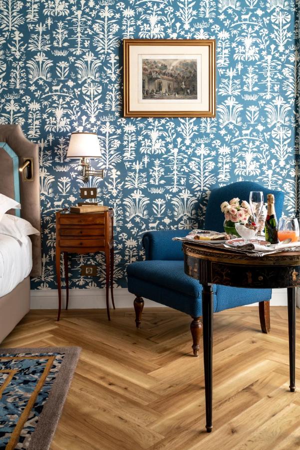 Where to Stay in Madrid: 14 Best Hotels 51 A stylish bedroom—perfect for those wondering where to stay in Madrid first time—features blue and white floral wallpaper, a cozy armchair, wooden nightstand with lamp, framed artwork, and a table set with wine and roses on herringbone wood floors.
