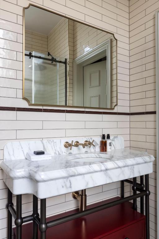 15 Best Boutique Hotels in London, UK 31 A bathroom inspired by boutique hotels in London, featuring cream subway tile walls, a rectangular mirror, and a white marble sink with gold fixtures. Toiletries rest on the sink, and a red cabinet is visible beneath the counter.