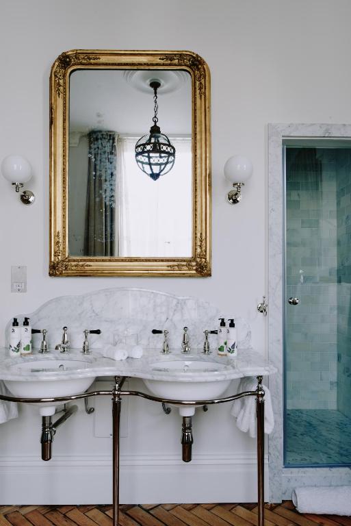 15 Best Boutique Hotels in London, UK 35 Elegant bathroom with a marble double sink, gold-framed mirror, wall sconces, and glass shower door—reminiscent of the luxurious style found in boutique hotels in London. Toiletries are neatly arranged, and a pendant light is reflected in the mirror.