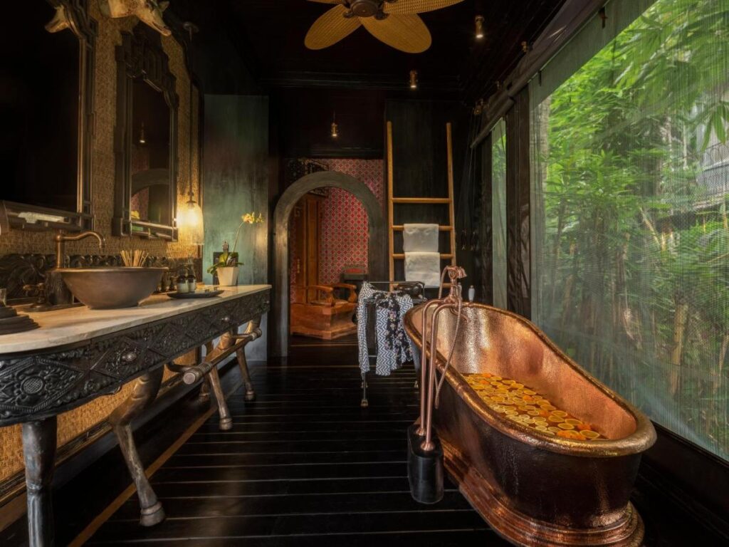 13 Most UNIQUE Places to Stay in Ubud 3 Luxurious bathroom with a copper bathtub filled with floating flower petals, ornate dark wood vanity, and a window overlooking lush bamboo—one of the unique places to stay in Ubud. Warm ambient lighting enhances the elegant decor.