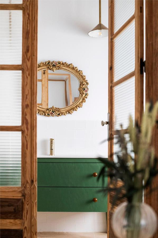 25 Weekend Trips from Madrid Max. 2h Away 71 A bathroom with a green vanity, gold faucet, and ornate gold-framed mirror, seen through wooden glass doors—reminiscent of stylish interiors found on weekend trips from Madrid. A pendant light glows above a blurred plant in the foreground.
