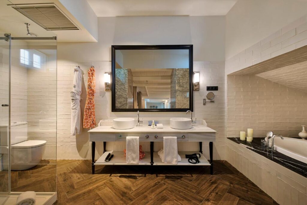Where to Stay in Madrid: 14 Best Hotels 45 Modern bathroom with double sinks, a large mirror, glass shower, white tiled walls, wooden herringbone floor, towels, candles by a bathtub, and bathrobes—ideal for those seeking where to stay in Madrid first time.