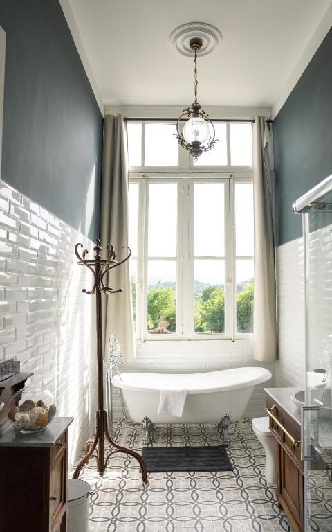 Bright bathroom inspired by boutique hotels in Asturias, featuring a large window, freestanding bathtub, patterned tile floor, white subway tile walls, vintage chandelier, wooden coat rack and vanity. Natural light fills the space.