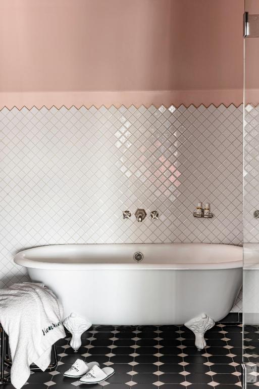 15 Best Boutique Hotels in London, UK 51 A white clawfoot bathtub stands against a wall with white diamond tiles and a pink upper section, reminiscent of the chic bathrooms found in boutique hotels in London. A towel and slippers rest on the black-and-white patterned floor next to the tub.