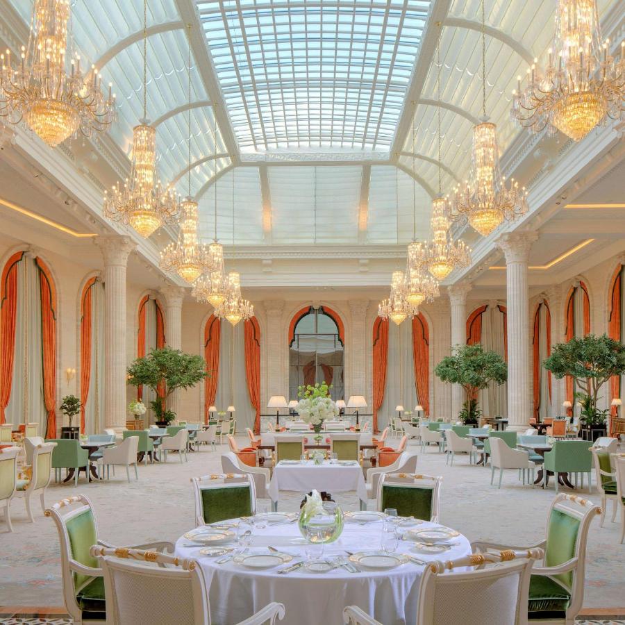 Where to Stay in Doha, Qatar: Transit, City & Beach Hotels 29 A grand, elegant dining hall—ideal for those exploring where to stay in Doha—with tall columns, lush green plants, high arched windows with orange drapes, crystal chandeliers, and a large glass ceiling; tables set in a bright, luxurious setting.
