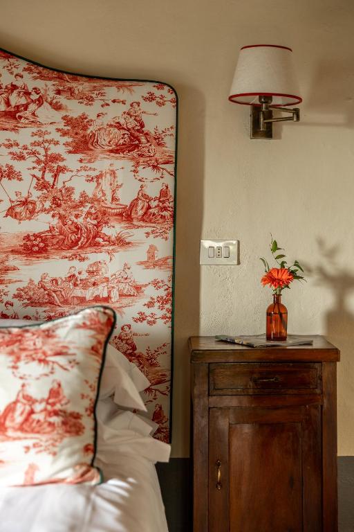 7 Best Agriturismos in Tuscany 28 A cozy bedroom corner inspired by agriturismos in Tuscany, featuring a wooden nightstand, an orange flower in a brown glass vase, a decorative headboard and pillow with red pastoral patterns, and a wall-mounted light above.