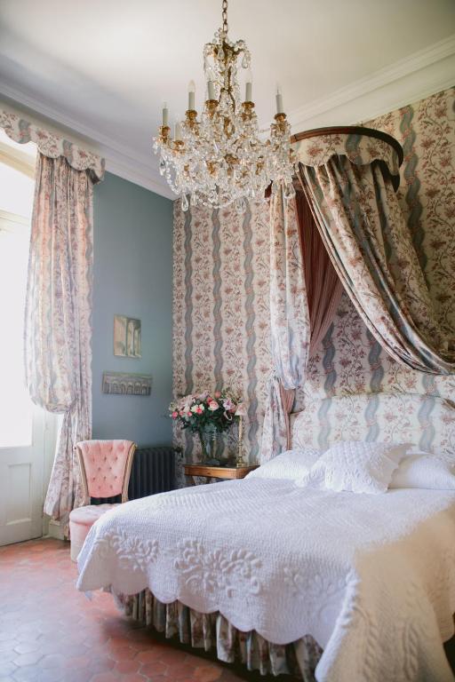 11 Unique Places to Stay in Provence 15 A vintage-style bedroom with ornate wallpaper, a canopy bed covered in a white quilt, a crystal chandelier, and floral curtains embodies the charm of unique places to stay in Provence, with roses on the table by the window.