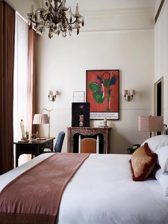 15 Best Boutique Hotels in London, UK 43 A stylish bedroom reminiscent of boutique hotels in London, with a neatly made bed, rust-colored throw and pillows. There’s a decorative fireplace, modern artwork above it, chandelier, desk with lamp, and tall windows with pinkish curtains.