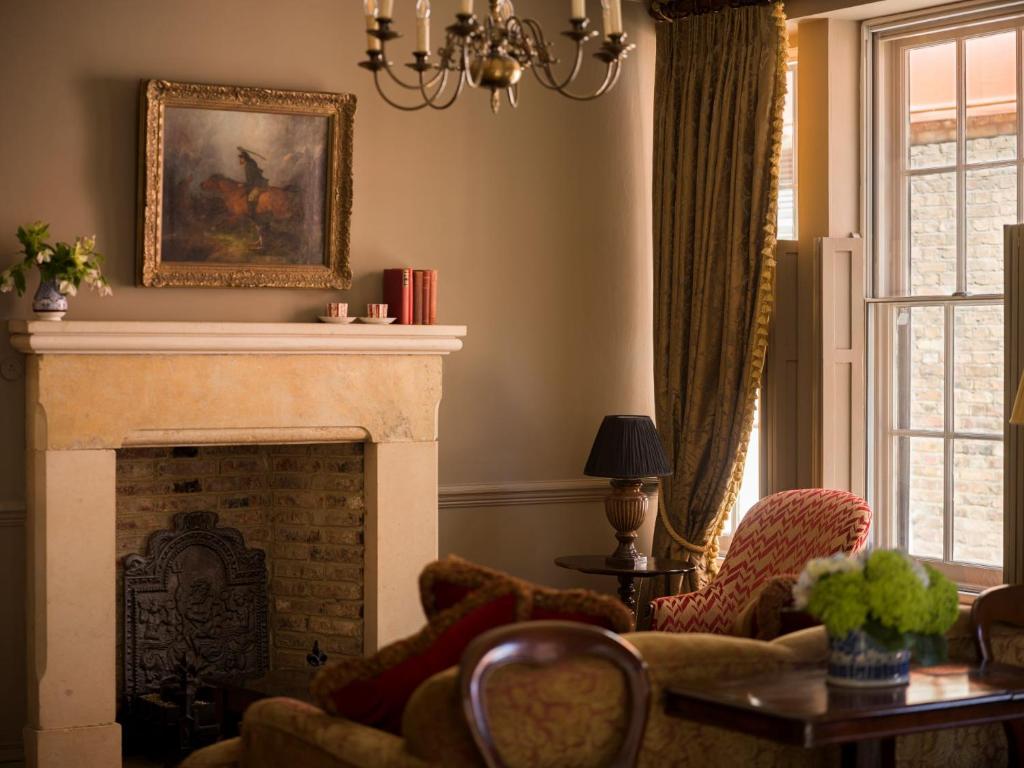 15 Best Boutique Hotels in London, UK 40 A cozy living room reminiscent of boutique hotels in London, featuring a stone fireplace, armchair, sofa, side table with a lamp, golden-curtained window, framed painting above the mantel, and a vase with flowers bathed in warm light.