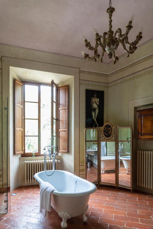 7 Best Agriturismos in Tuscany 22 A vintage bathroom reminiscent of charming agriturismos in Tuscany, featuring a freestanding clawfoot bathtub, a towel draped over the edge, a large window with wooden shutters, a chandelier, and a folding mirror with a painting on the wall.