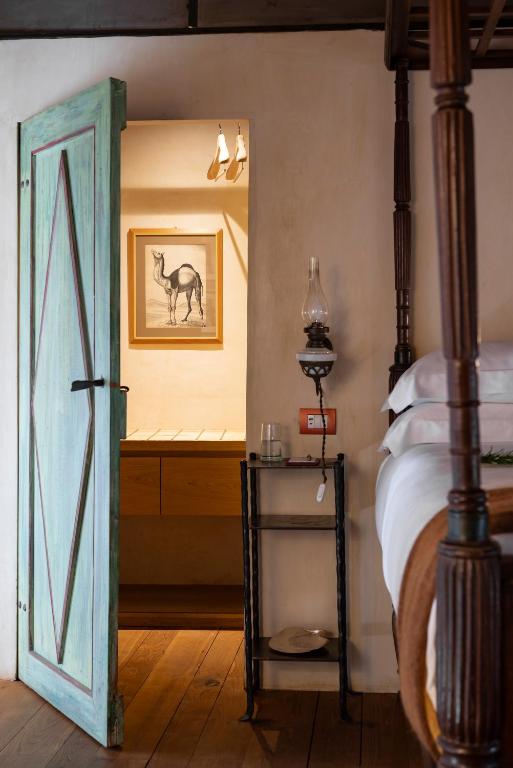 7 Best Agriturismos in Tuscany 21 A rustic bedroom reminiscent of agriturismos in Tuscany features a turquoise wooden door, slightly open to a bathroom with a framed camel print. Wooden floors, a vintage lamp, and a small bedside shelf with glass and book complete the cozy scene.