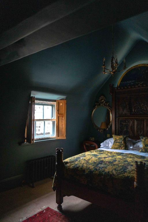 15 Best Boutique Hotels in London, UK 41 A dimly lit bedroom with dark green walls, a wooden bed with yellow floral bedding, an ornate headboard, and an antique-style chandelier evokes the charm of boutique hotels in London, with a small window and elegant mirror completing the scene.