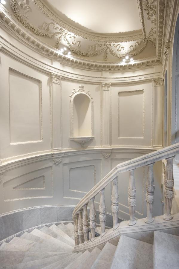 10 UNIQUE Hotels with Pool in Seville 16 A curved marble staircase with ornate balustrades ascends beside cream-colored, intricately decorated walls—evoking the elegant, classical interiors found in some of the finest hotels with pool in Seville. Soft lighting highlights every refined detail.