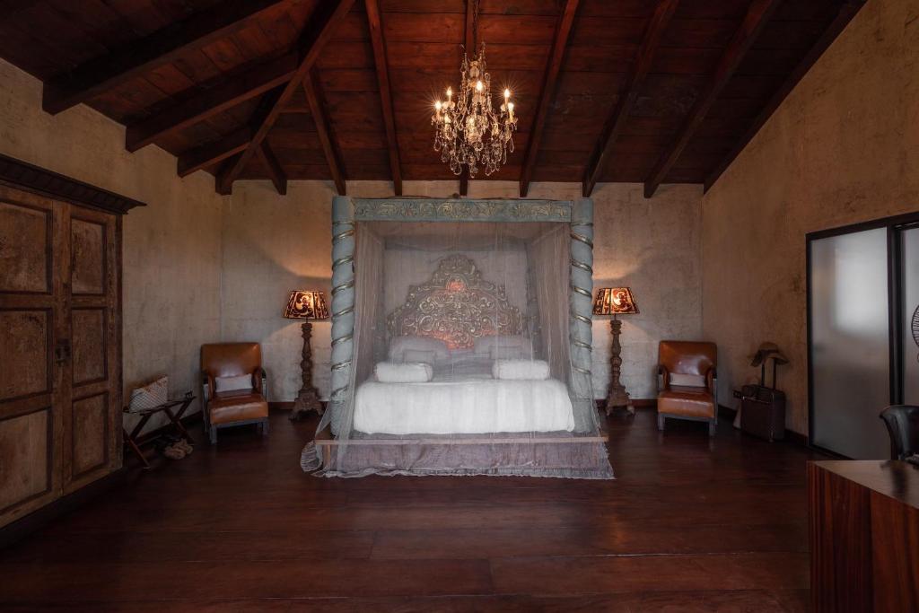8 UNIQUE Places Where to Stay in Lake Atitlán 4 A rustic bedroom with a high wooden ceiling, chandelier, large ornate canopy bed, leather armchairs, vintage lamps, and muted earthy tones—perfect inspiration if you’re searching where to stay in Lake Atitlan.