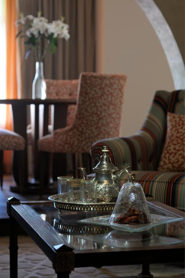 Where to Stay in Doha, Qatar: Transit, City & Beach Hotels 39 A glass coffee table with a silver tea set and dates sits in the foreground, while patterned chairs and a round table suggest where to stay in Doha. A vase of white flowers and warm lighting fill the cozy, inviting room.