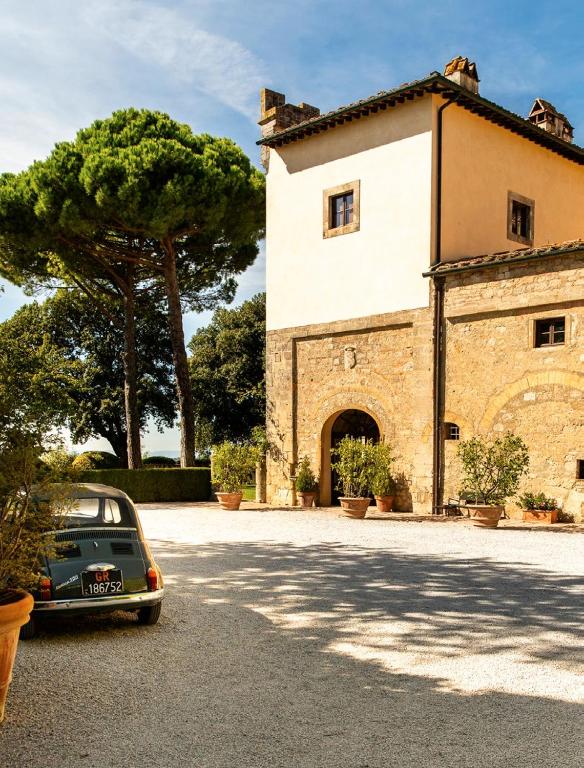 7 Best Agriturismos in Tuscany 25 A vintage car is parked on a gravel driveway beside a rustic stone villa—reminiscent of agriturismos in Tuscany—surrounded by potted plants, large green trees, and a clear blue sky in the background.