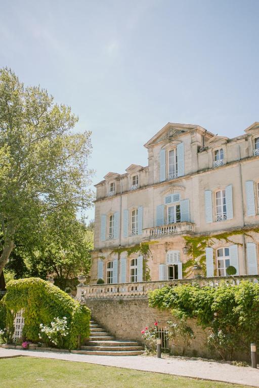 11 Unique Places to Stay in Provence 14 A large, elegant stone house with blue shutters and green ivy climbing its walls, surrounded by trees and flowering bushes under a clear sky—one of the most unique places to stay in Provence. Steps lead up to the welcoming main entrance.