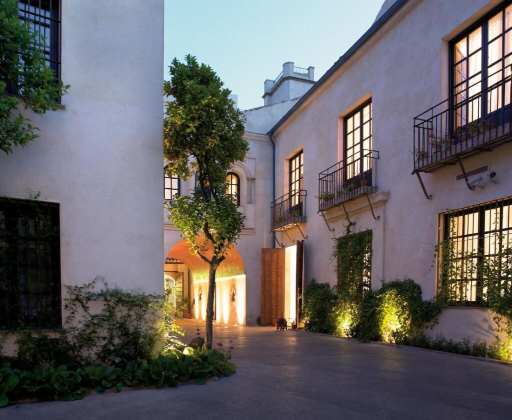 Best of Cordoba One Day Itinerary 4 A serene courtyard in Cordoba, perfect for a one day visit, features a single tree, climbing plants, and warm lighting along white stucco buildings with balconies and softly illuminated windows at dusk.