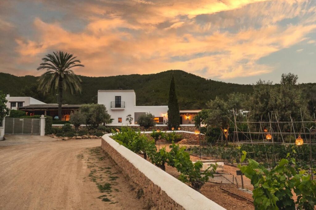 10 AUTHENTIC Finca Hotels in Ibiza 29 A Mediterranean-style villa with white walls sits amid lush greenery at sunset, warm lights glowing as mountains rise behind. This tranquil scene is reminiscent of finca hotels in Ibiza, blending natural beauty with relaxed luxury.