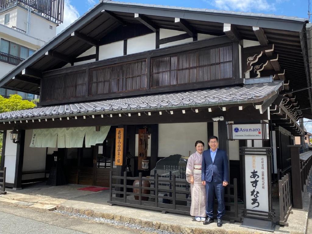 Looking for the Best Ryokan in Takayama? 10 Two people stand smiling in front of a traditional Japanese wooden building with a tiled roof, known as the Best Ryokan in Takayama. One wears a kimono and the other a suit. Signs beside them display Japanese text and the word Asunaro.
