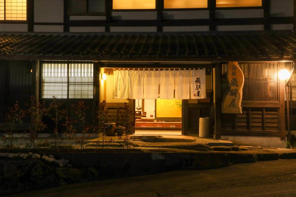 Looking for the Best Ryokan in Takayama? 3 A warmly lit traditional Japanese inn entrance at night showcases wooden architecture, paper lanterns, a noren curtain, and plants beside the stone path—inviting you to experience the Best Ryokan in Takayama.