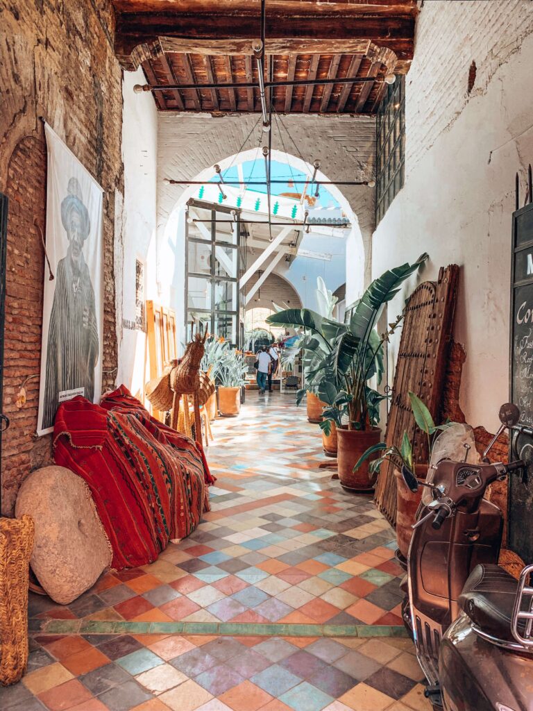 12 Charming Budget Riads in Marrakech 1 A sunlit hallway in one of the budget riads in Marrakech features a colorful tiled floor, potted plants, and rustic decor with woven fabrics and artwork. People are visible in the distance beneath an arched glass ceiling.