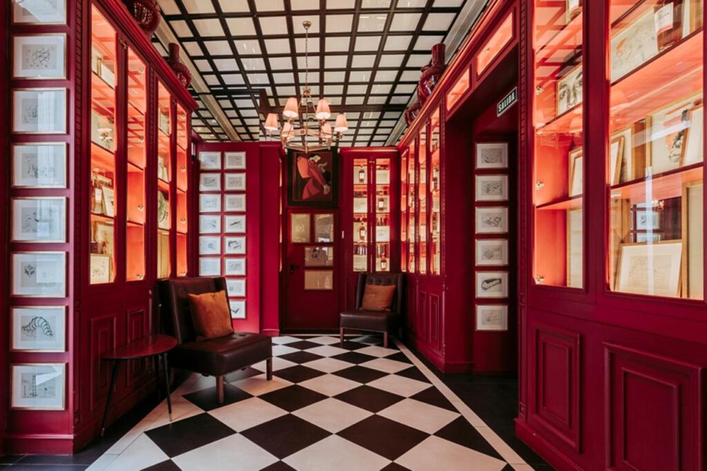Where to Stay in Madrid: 14 Best Hotels 47 A stylish hallway, perfect for those exploring where to stay in Madrid first time, with red walls, illuminated display shelves of books and art, black armchairs with cushions, a checkered tile floor, and a modern chandelier hanging from a grid-patterned ceiling.