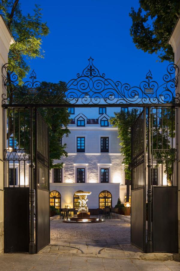 Where to Stay in Madrid: 14 Best Hotels 19 Ornate iron gates open onto a courtyard with a central fountain, leading to an elegant multi-story building—an inviting choice for those wondering where to stay in Madrid first time, framed by tall trees at dusk.