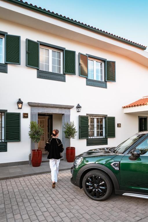 11 UNIQUE Boutique Hotels in Tenerife 42 A person walks toward the entrance of a white house with green shutters and potted plants, reminiscent of the charm found at boutique hotels in Tenerife. A green car is parked on the paved driveway in front.