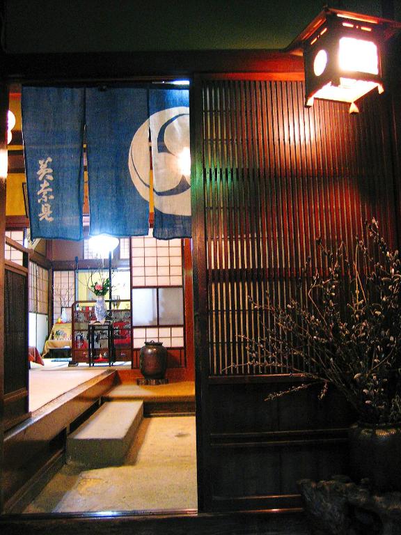 Looking for the Best Ryokan in Takayama? 7 A warm, inviting entrance to a traditional Japanese room at the best ryokan in Takayama features wooden sliding doors, a blue noren curtain with white patterns, tatami mats, and soft lantern lighting. A vase with branches decorates the entryway.