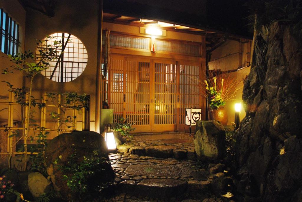 7 UNIQUE Ryokans in Gion Kyoto 15 A warmly lit traditional Japanese entrance, like those found at ryokans in Gion Kyoto, features wooden sliding doors, a round window, stone pathway, and lush plants, creating an inviting and serene atmosphere at night.
