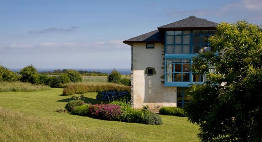 A modern house with blue window frames stands surrounded by green grass and trees, offering a distant sea view and partly cloudy sky—reminiscent of the charming boutique hotels in Asturias.