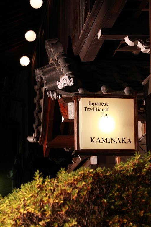 Looking for the Best Ryokan in Takayama? 18 A warmly lit sign for Japanese Traditional Inn KAMINAKA, known as one of the Best Ryokan in Takayama, hangs under a traditional tiled roof at night, with glowing lanterns above and green foliage in the foreground.